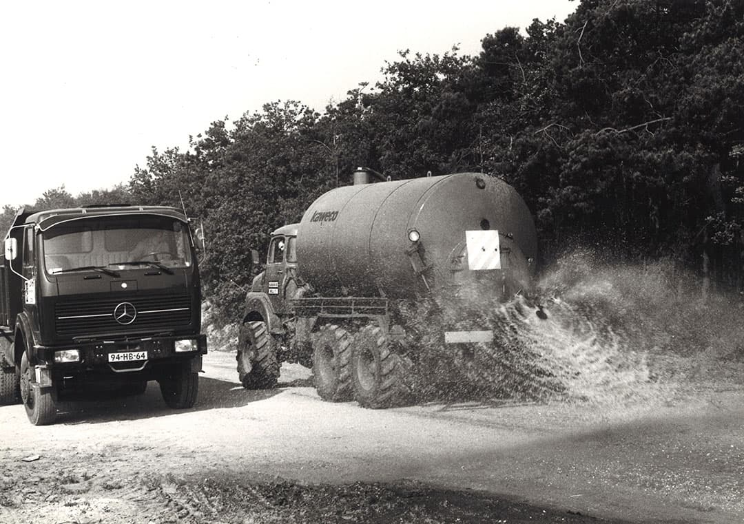 Zandwegen waren vroeger heel gewoon op het platteland. Ze waren niet ideaal. Bij nat weer werden het moddergeulen, bij droogte mulle zandbakken. Bovendien zorgde het stuiven 's zomers voor overlast. Op de foto werd gesproeid om dat tegen te gaan. Deze aanpak was geen blijvertje. - Foto: Misset